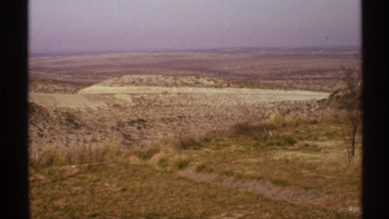 Dry New Mexico landscape viewed from above with a dirt path, scrub grass, rolling hills, and hazy sky.