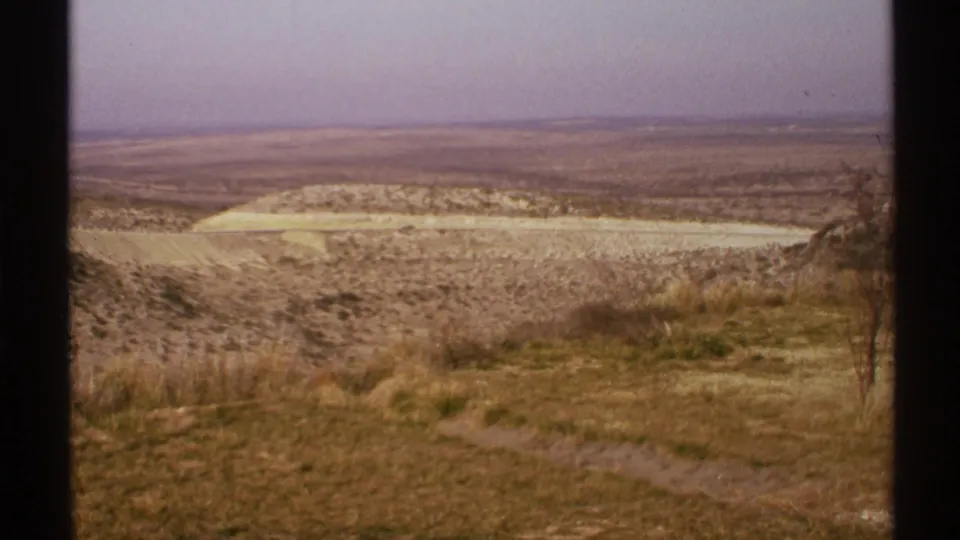 1972 8mm home-movie frame of an arid New Mexico overlook with dry grass, a faint path, pale hills, and a broad hazy horizon.