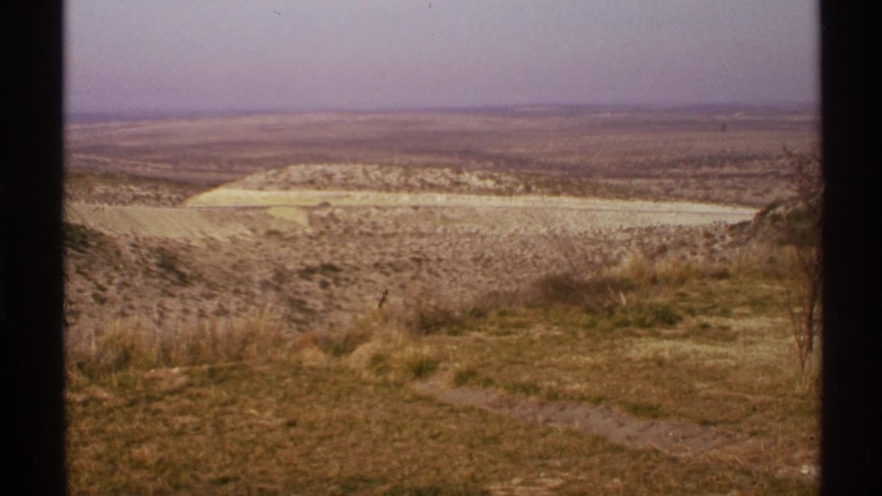 Open arid plain with dry foreground grass, low hills, pale ground, and a hazy horizon.