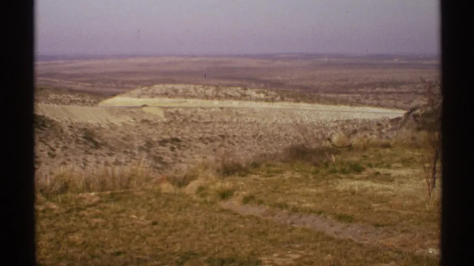 1972 8mm frame of a New Mexico arid overlook, showing dry foreground vegetation, a narrow path, pale hills, and distant flat land.