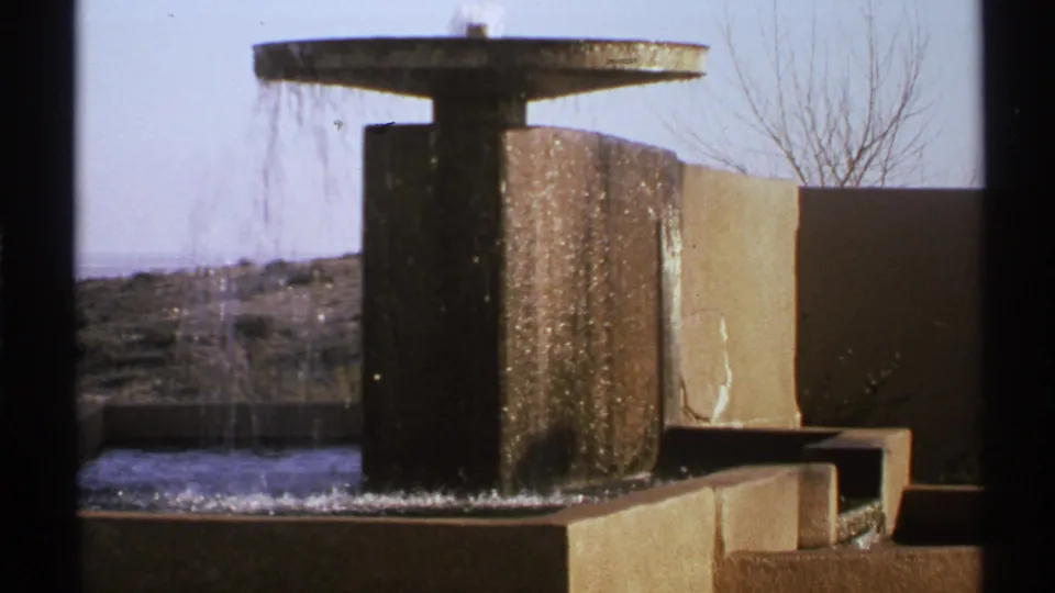 1972 New Mexico 8mm frame showing a modern concrete fountain with flowing water, low walls, distant landscape, and a leafless tree.