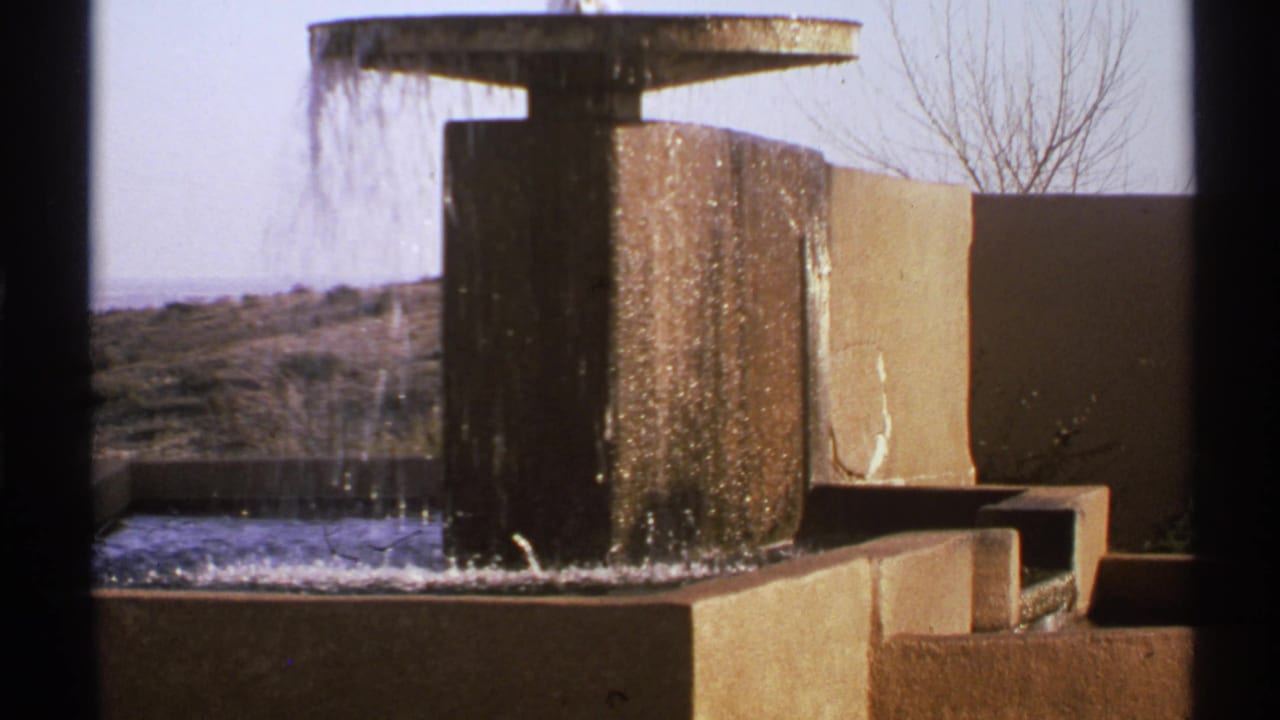 1972 New Mexico home-movie frame of a concrete fountain, angular walls, bare branches, and pale sky.