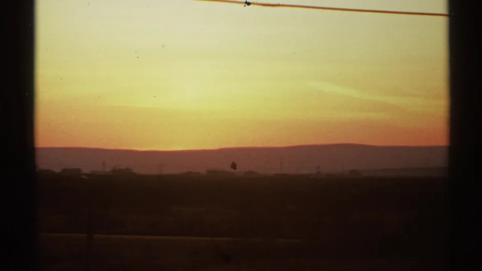 Golden New Mexico sunset over a low horizon, seen through dark window or vehicle framing on 8mm film.