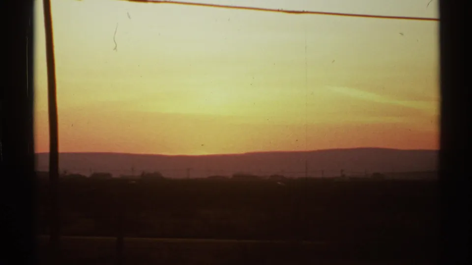 New Mexico sunset on 8mm film with a pale golden sky, distant low hills, dark terrain, and vertical window framing.