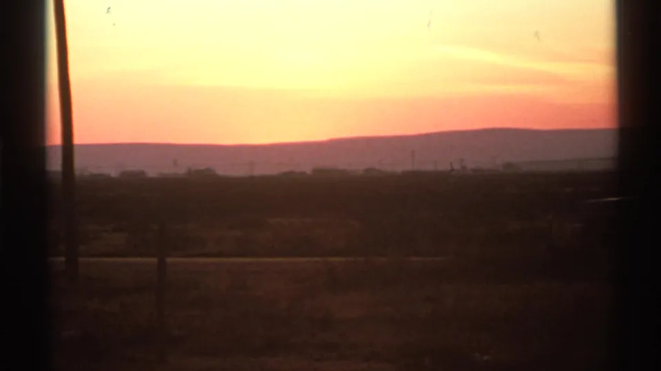 1972 home-movie frame of a New Mexico sunrise with open terrain, distant hills, faint utility features, and visible 8mm grain.