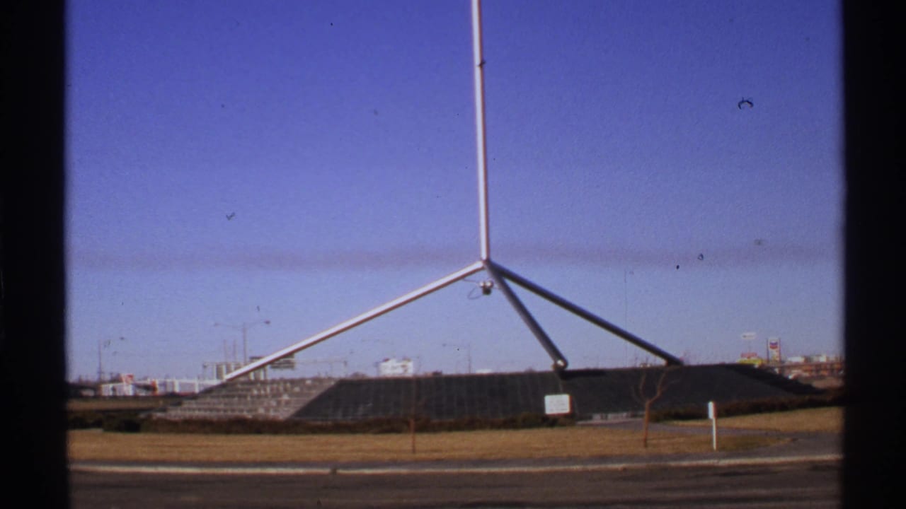 Silver tripod-like metal sculpture beside a road under blue sky.