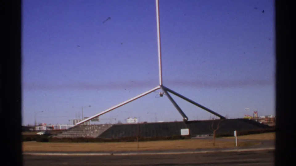 1972 8mm view of a silver tripod-like sculpture near a road, with distant buildings and visible dark frame edges.