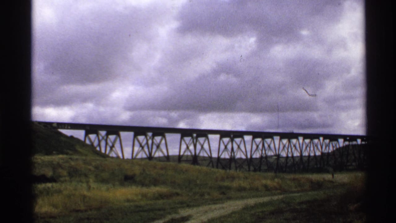 1972 North Dakota 8mm frame of a long trestle bridge crossing open grassland beneath heavy clouds, viewed through a dark window frame.