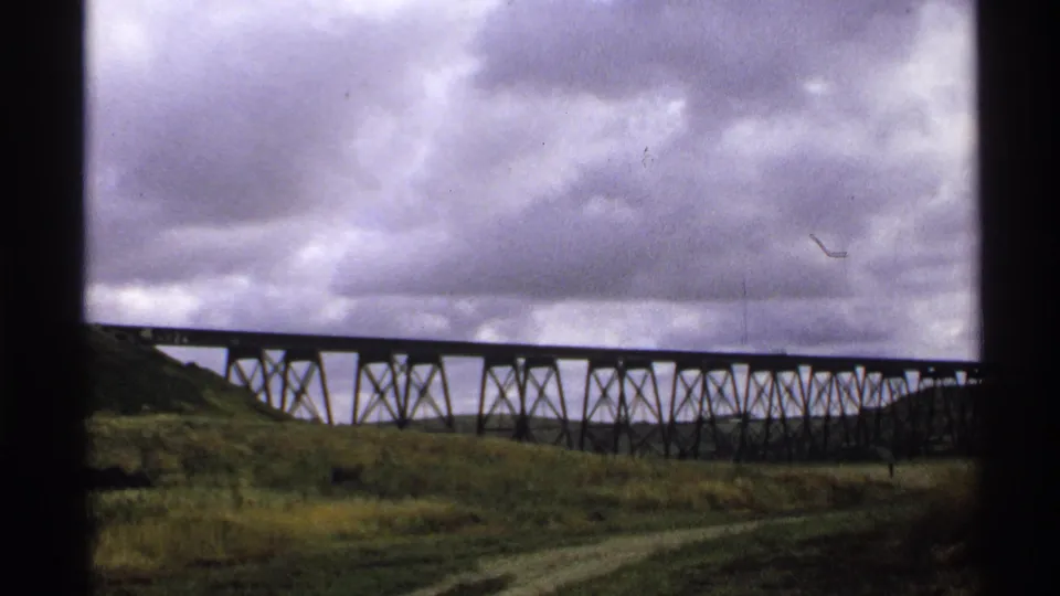 1972 North Dakota 8mm frame of a long trestle bridge crossing open grassland beneath heavy clouds, viewed through a dark window frame.