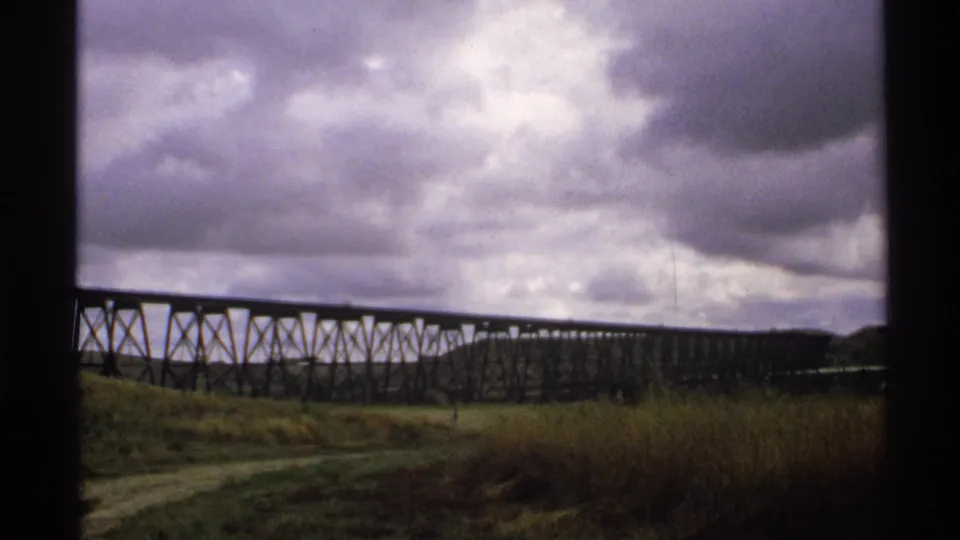 8mm frame showing the long trestle bridge and its repeating support pattern above rough grass and a pale dirt track.