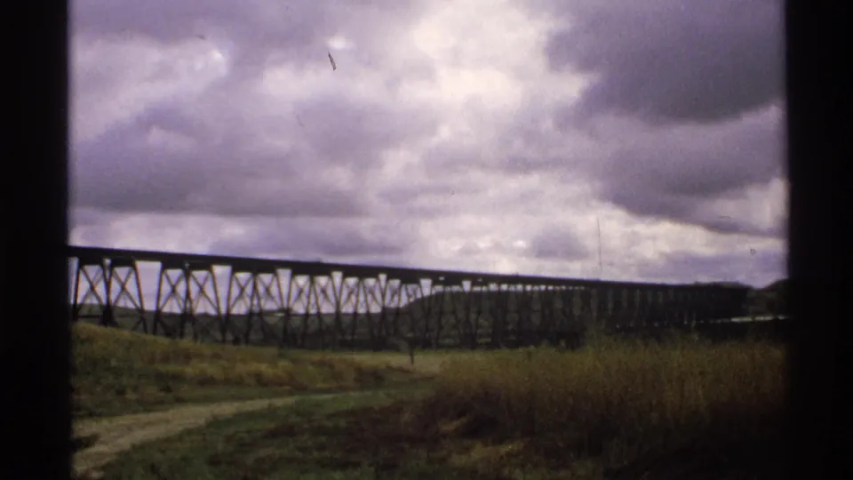 1972 North Dakota 8mm frame showing a long trestle bridge across grassland under a dark, cloud-filled sky.