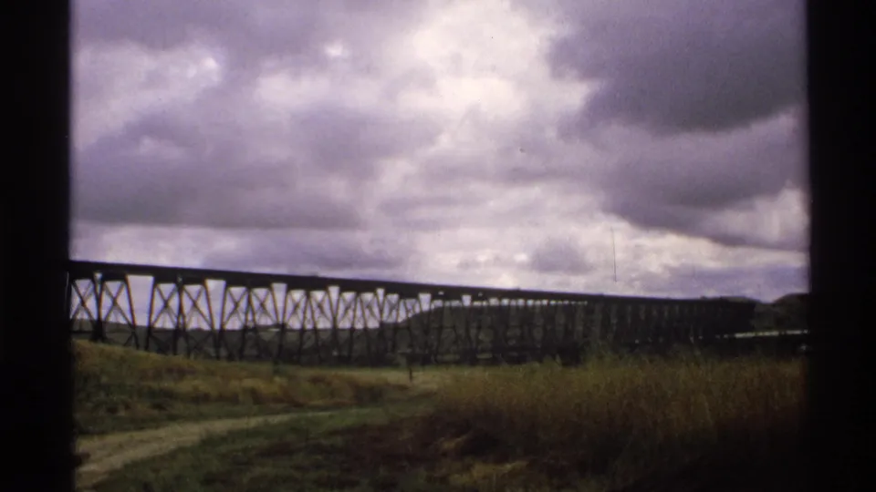 1972 North Dakota home-movie view of a dark trestle bridge, roadside grass, and low stormy clouds.