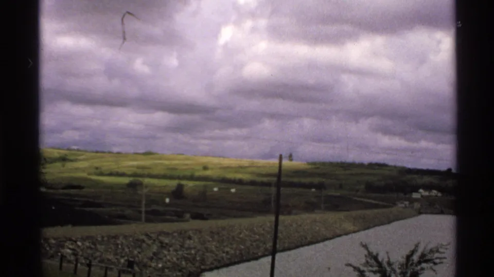 1972 North Dakota 8mm landscape with rolling hills, a pale road, utility poles, and a distant waterway under overcast clouds.
