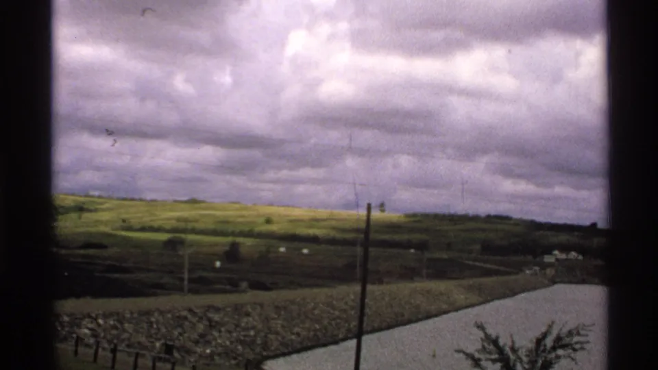 1972 North Dakota frame showing a rural road, open hills, fence lines, and heavy purple-gray clouds.