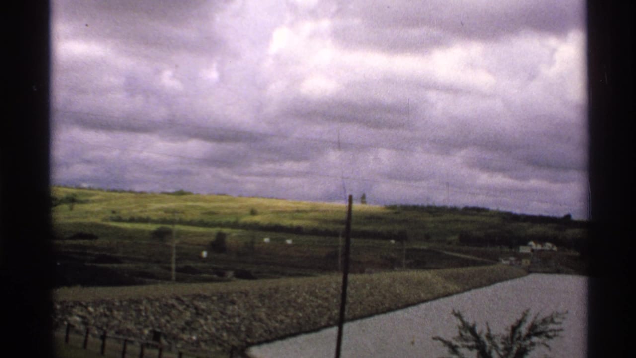 1972 North Dakota rural view through a window, showing green fields, utility poles, a cloudy sky, and a stone-lined pond or reservoir.