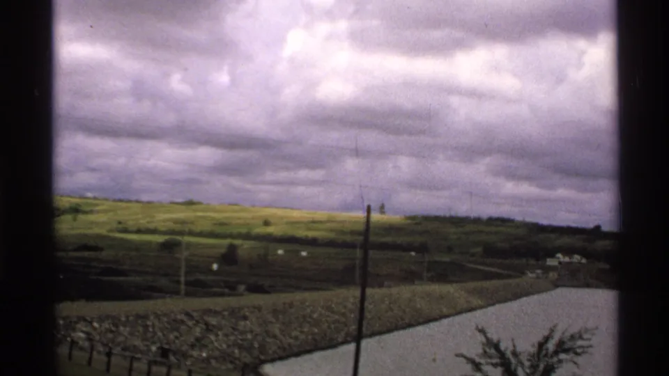1972 North Dakota rural view through a window, showing green fields, utility poles, a cloudy sky, and a stone-lined pond or reservoir.
