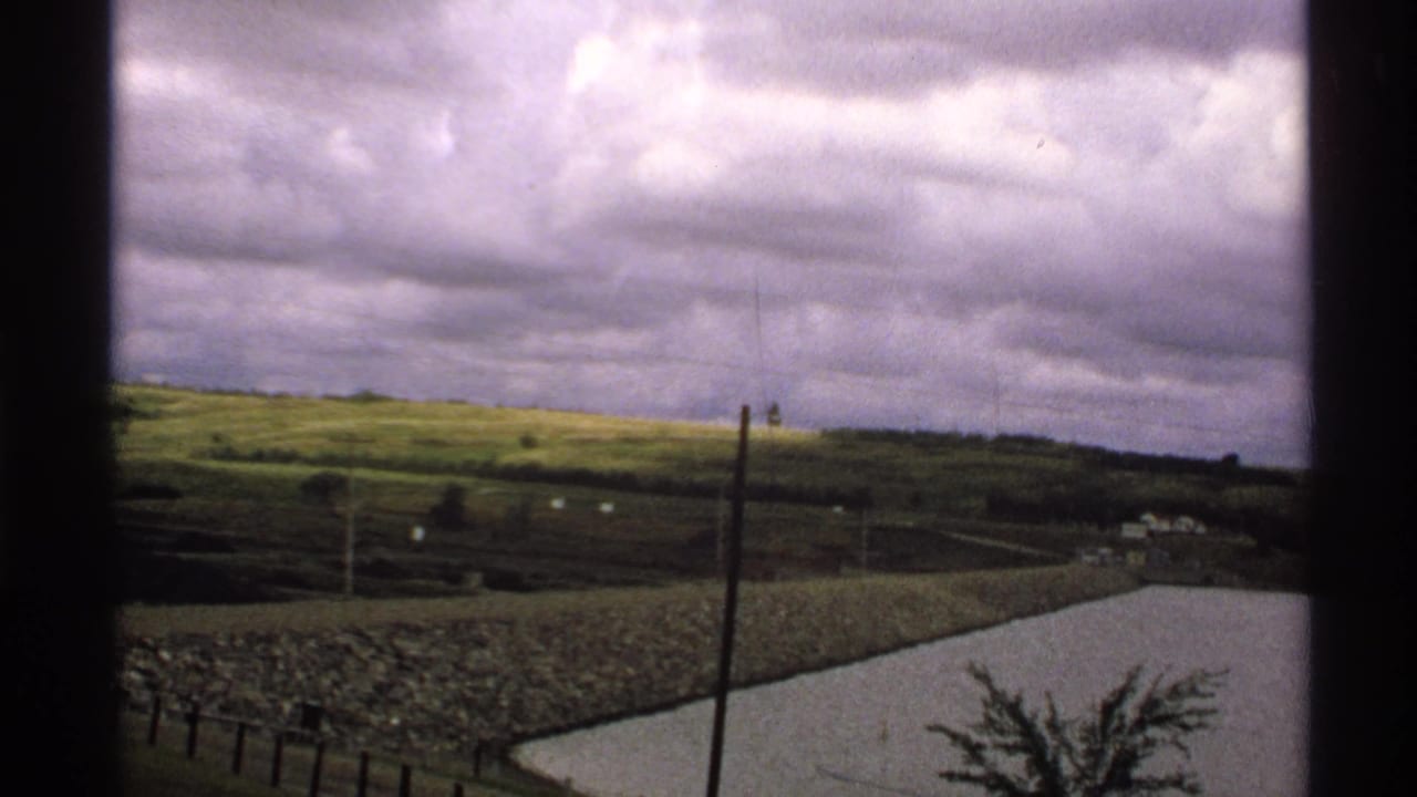 Rural North Dakota countryside viewed through a window, with green hills, utility poles, distant structures, and a pale pond or reservoir.