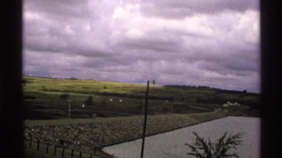 Rural North Dakota countryside viewed through a window, with green hills, utility poles, distant structures, and a pale pond or reservoir.