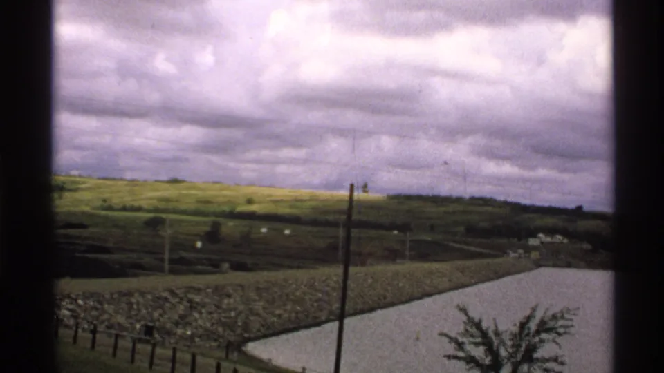 1972 rural North Dakota view framed by dark window bars, showing fields, fencing, utility poles, and a stone-edged pond or reservoir.