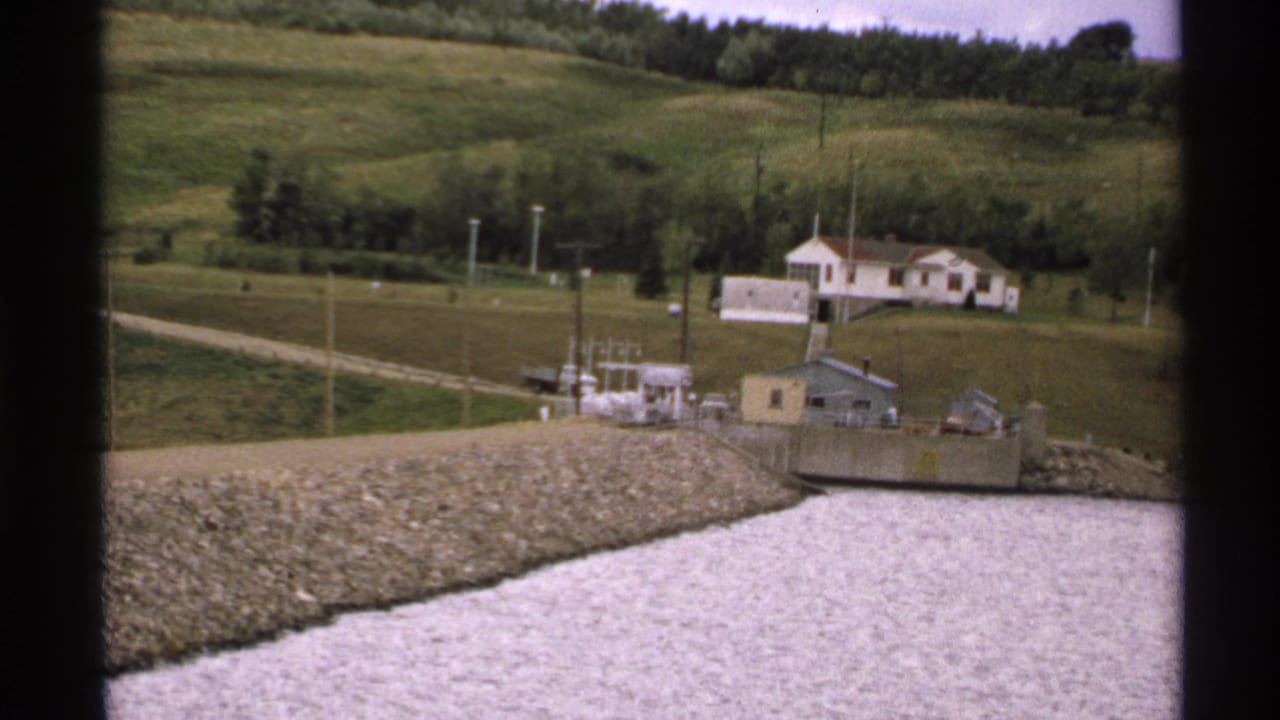 1972 North Dakota rural waterside view with riprap embankment, water-control structure, utility poles, and a white house on a grassy hill.