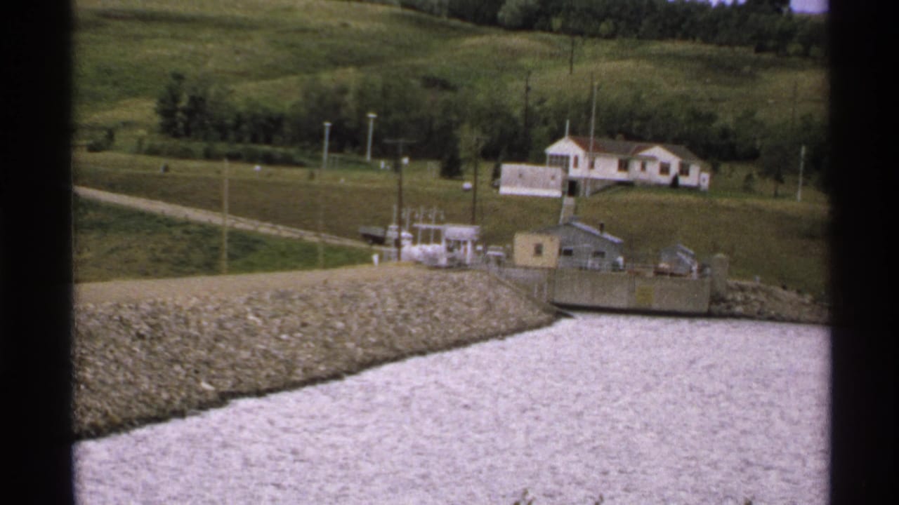 North Dakota home-movie frame showing a calm river edge, riprap slope, small utility or dam equipment, dirt road, and hillside house.
