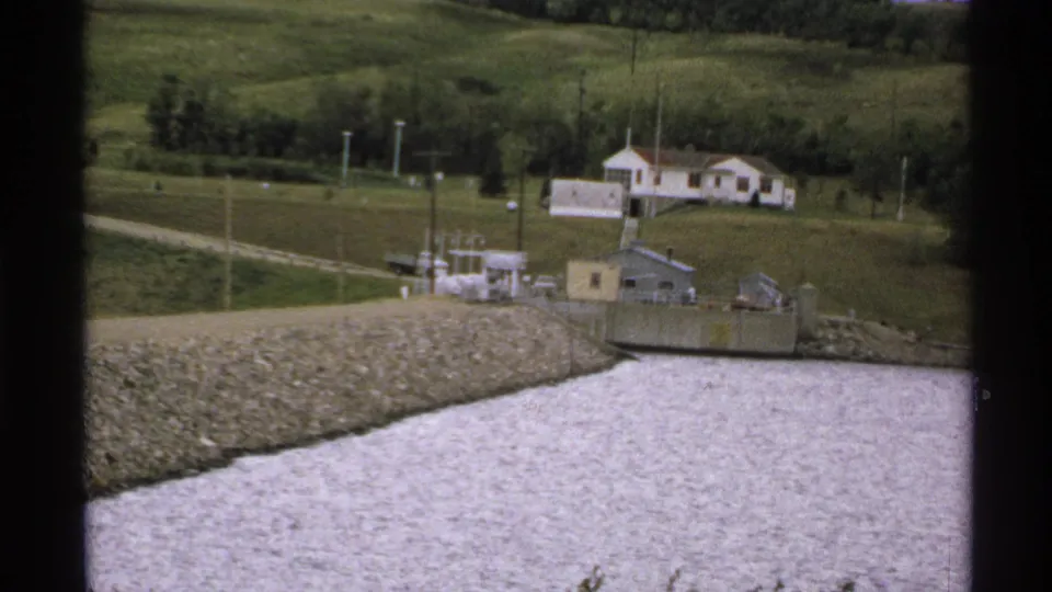 Muted 8mm frame of a North Dakota river or reservoir beside a gravel embankment, with small utility structures and a rural house in the background.