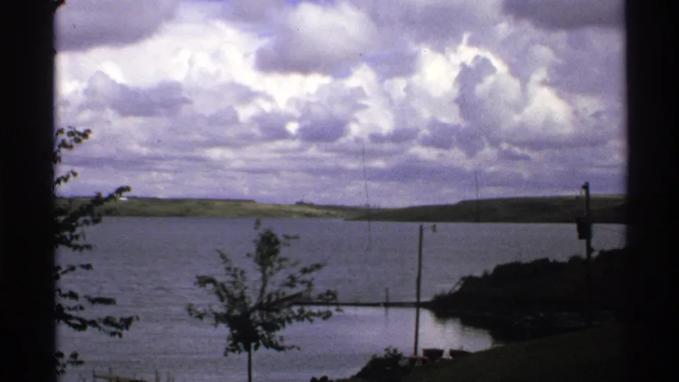 1972 North Dakota waterside landscape with broad cloudy sky, distant shoreline, calm water, poles, trees, and dark window-like framing.