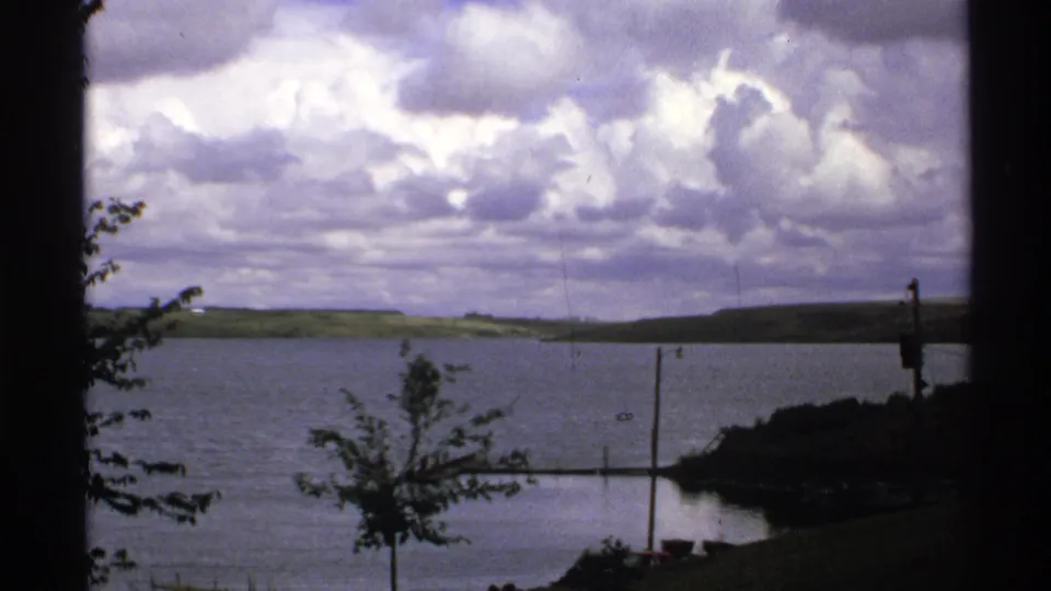 1972 North Dakota 8mm frame showing a calm lake below heavy clouds, with distant hills, a young tree, dock sections, and dark film edges.
