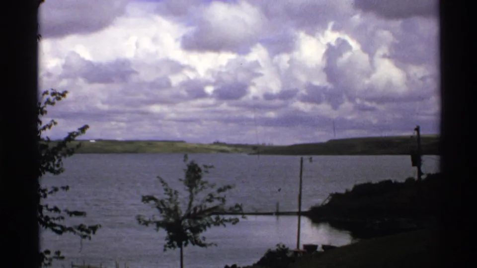 1972 North Dakota 8mm frame of a still lake and low green hills, with a foreground tree, dock pieces, poles, and dramatic cloud cover.