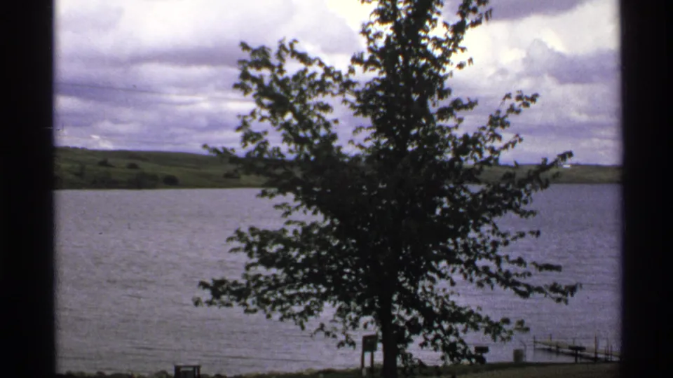 1972 North Dakota 8mm frame with a leafy tree centered before a calm lake, distant hills, cloud cover, and small waterfront structures.