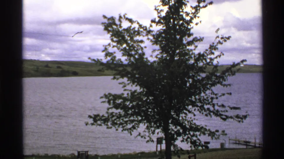 1972 North Dakota 8mm frame showing a foreground lakeside tree, broad water, low hills, dock railings, and textured cloud cover.
