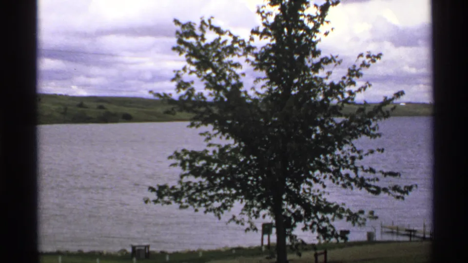 1972 North Dakota 8mm view of a leafy tree by a lake, with distant green hills, cloudy sky, and dark window-like framing.
