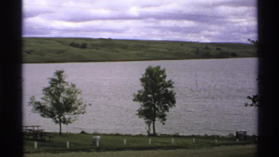1972 North Dakota 8mm frame showing two shoreline trees, lake water, picnic table, grassy bank, cloudy sky, and rolling hills.