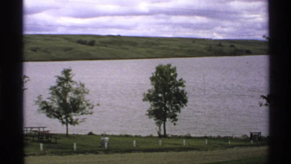 1972 North Dakota home-movie view of a grassy lakeside area with two trees, picnic furniture, white posts, lake, and distant hills.