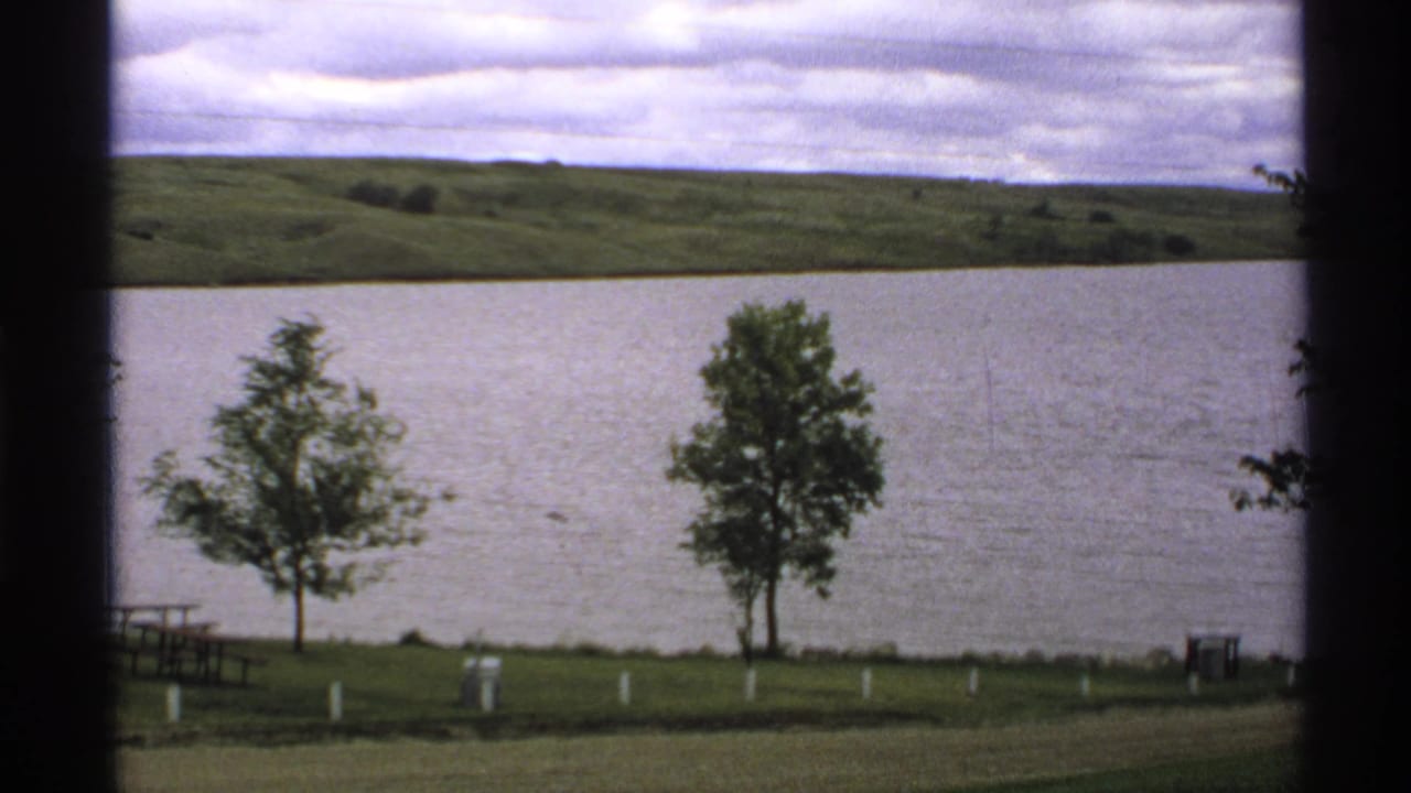 Lakeside park with two small trees, picnic tables, white posts, water, hills, and cloudy sky.