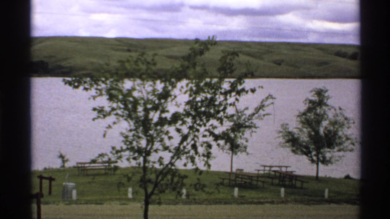 Tree branches in front of water, hills, picnic tables, white posts, grass, and cloudy sky.