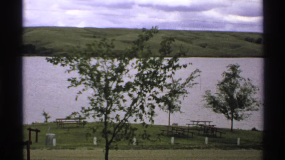 1972 North Dakota home-movie frame with leafy foreground branches, a lakeside picnic area, water, white posts, and green hills beyond.