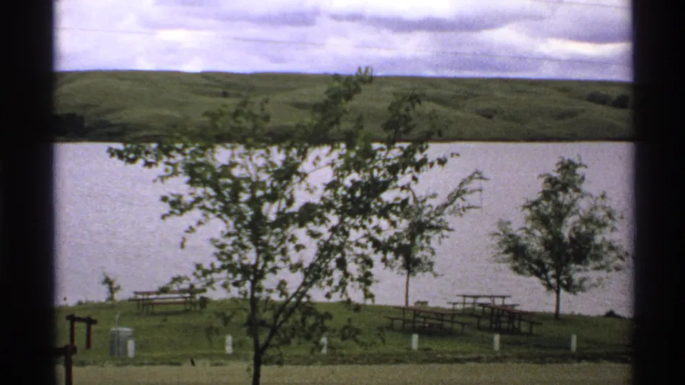 1972 North Dakota 8mm view of a leafy tree over a grassy shore with picnic tables, white posts, water, and rolling hills.