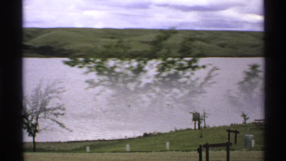 1972 North Dakota home-movie frame showing water and rolling hills, with a motion-blurred tree, grassy shoreline posts, and a small signboard.