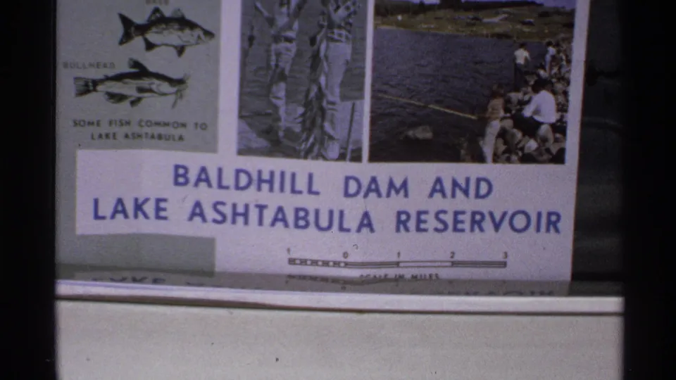 1972 North Dakota home-movie frame showing the reservoir sign with fish illustrations, fishing photo, and scale markings.