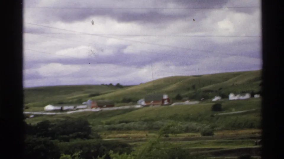 1972 North Dakota 8mm frame showing green rural hills, farm buildings, and a cloudy sky after the reservoir sign shot.