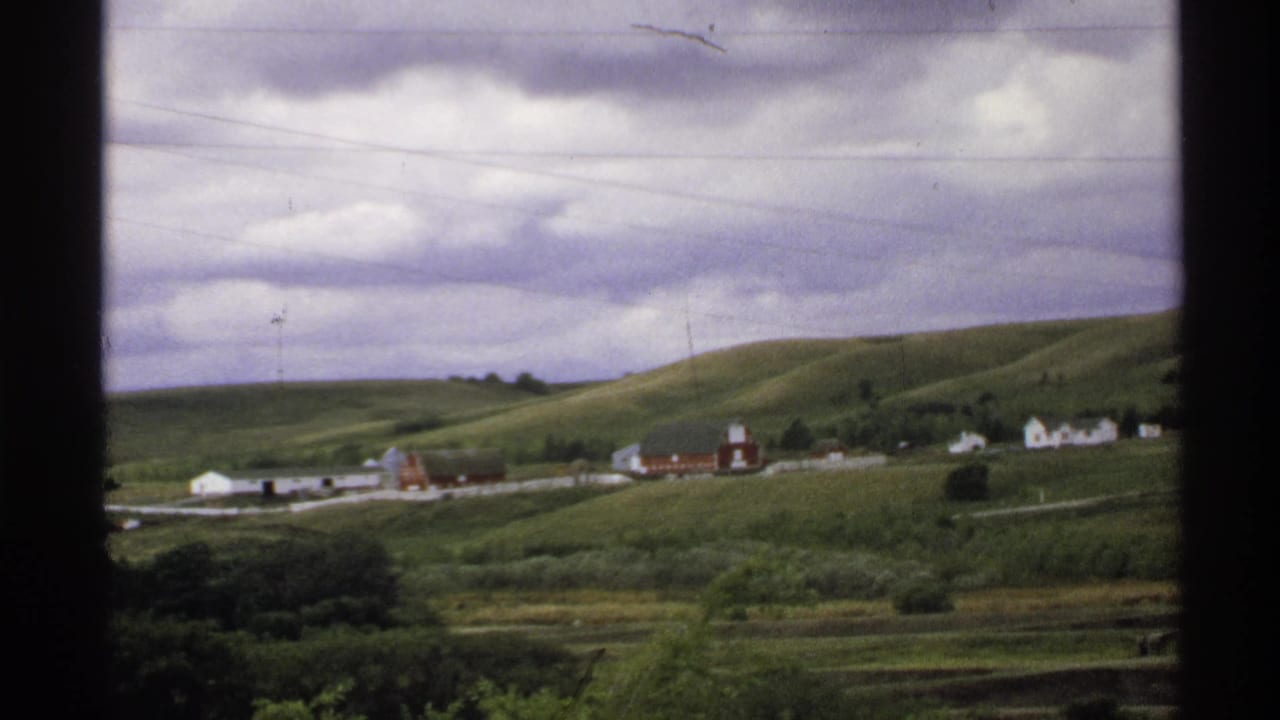 Rolling green hills with a distant farmstead, red barns, white buildings, power lines, and cloudy sky.