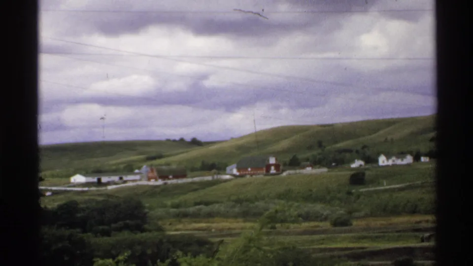 1972 North Dakota 8mm frame showing rolling hills, a distant farmstead, utility lines, and heavy clouds seen through dark frame edges.