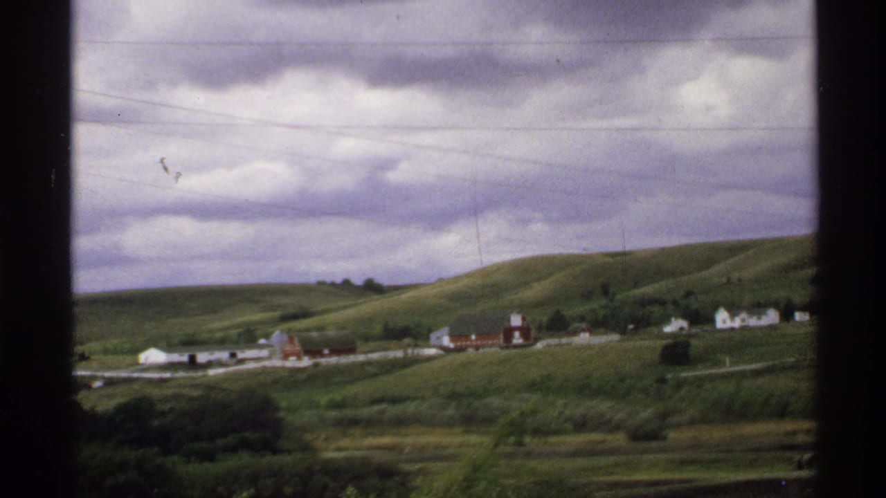 Distant farmstead on green hills with red barns, white buildings, power lines, and overcast clouds.