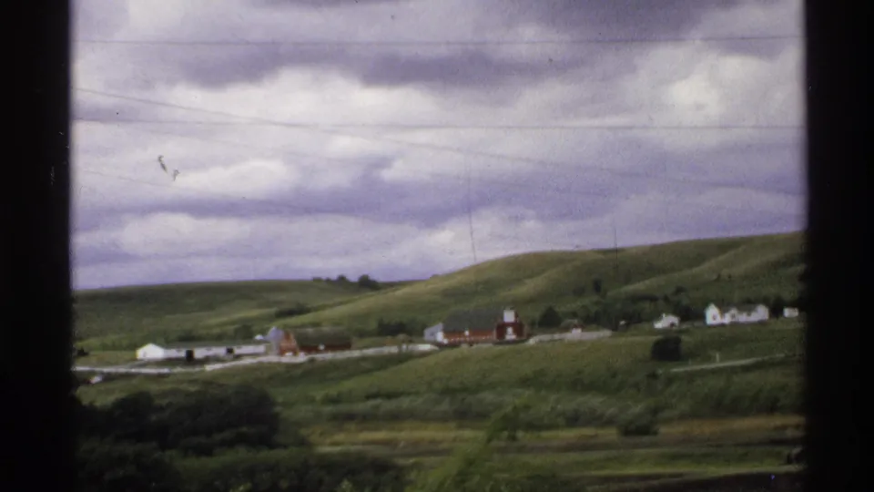 1972 North Dakota rural view with red barns and white buildings set into green hills beneath a layered cloudy sky.