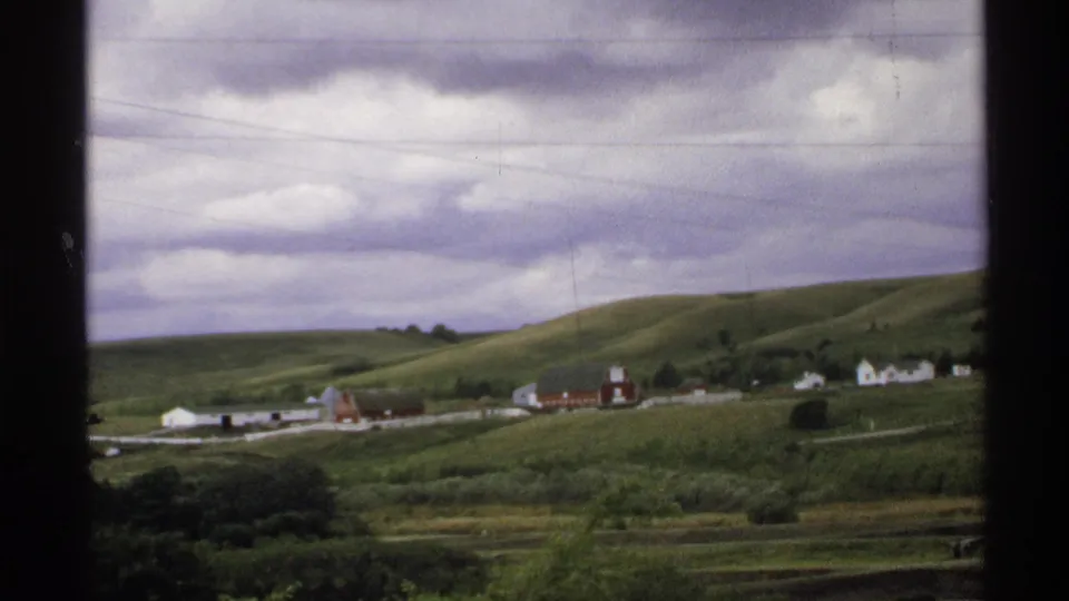 1972 North Dakota 8mm frame of a broad rural landscape with farm buildings, green slopes, power lines, and soft film grain.