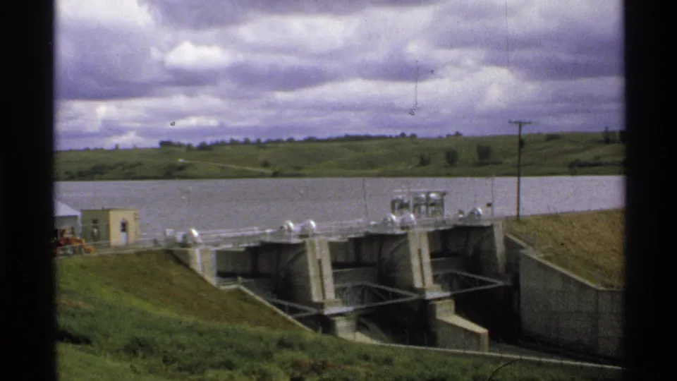 1972 North Dakota frame showing a reservoir and concrete dam infrastructure with grassy banks, utility poles, and distant rolling hills.