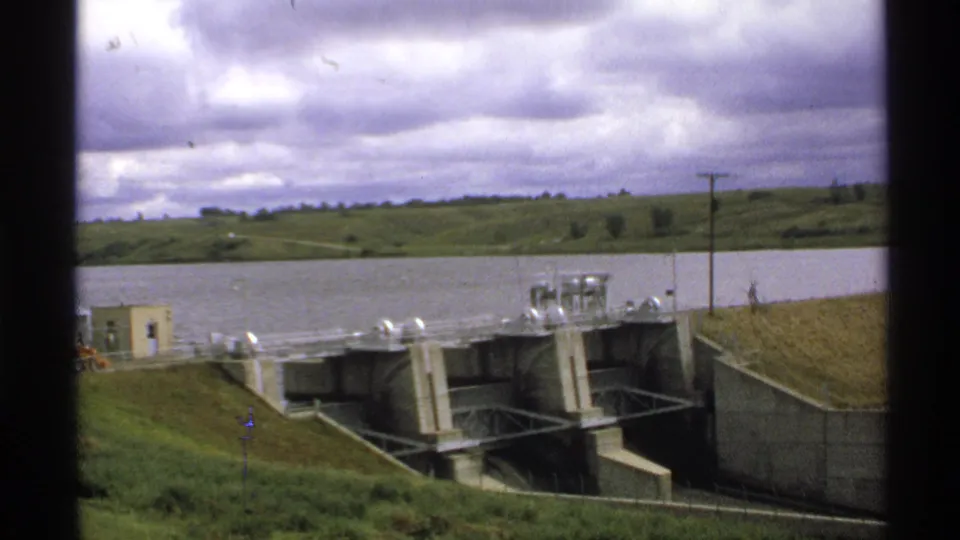 North Dakota 1972 8mm frame showing Baldhill Dam works, reservoir water, grassy banks, and cloudy sky.
