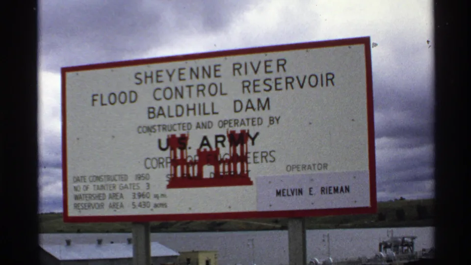North Dakota 1972 frame of the Baldhill Dam sign, with reservoir water, low buildings, and cloudy sky behind.