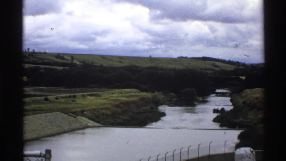 1972 North Dakota home-movie frame showing the reservoir outflow or river corridor near Baldhill Dam beneath a cloudy sky.